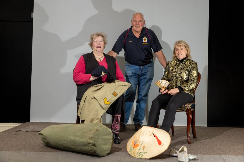 Minefields and miniskirts cast members Trish Storr and Rosie Knight sitting on chairs on a stage with Terang RSL sub-branch president Terry Fidge standing in the centre with items representing the Vietnam War. Picture by Eddie Guerrero